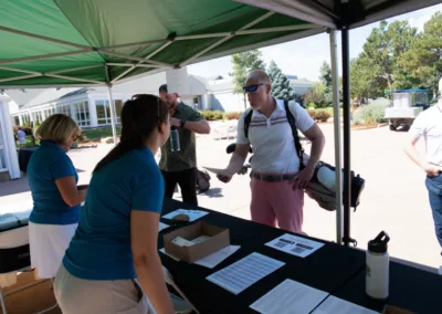 Several people are gathered under a green canopy tent at an outdoor event. A man with a golf bag is shaking hands with a woman behind a registration table containing papers and name tags. Other participants are nearby; trees and buildings are visible in the background.