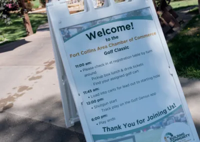 A close-up of an event sign on a pathway. The sign welcomes participants to the Fort Collins Area Chamber of Commerce Golf Classic and provides a schedule for the day, including registration, lunch pickup, golf start time, tracking on an app, and end time.