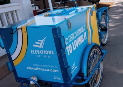 A blue utility bike with a large cargo box displaying advertisements for Elevations Credit Union. The box features the slogan "Go from dreaming it to living it" and includes the website elevationscu.com. The bike is parked on a sidewalk near a white fence.