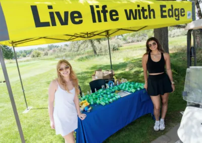 Two women are standing under a yellow canopy tent with the text "Live life with Edge." The tent covers a table filled with assorted items, possibly event giveaways. Both women are casually dressed and smiling. The backdrop includes trees and grass, suggesting an outdoor event.