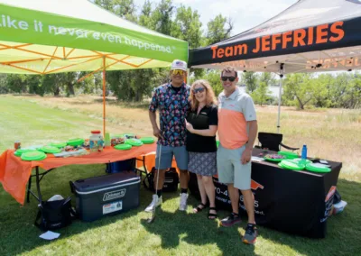 Three people are standing together, smiling, on a grassy area at a golf event. They are under bright tents with texts "Like it never even happened." and "Team JEFFRIES" on them. There are tables with food and drinks, and trees are visible in the background.