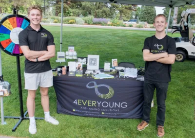 Two smiling people stand arm-in-arm in front of a booth with a banner reading "4EVERYOUNG Anti Aging Solutions." The table displays various products, while a colorful wheel is to the side. The outdoor setting includes grass and trees in the background.