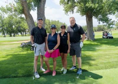 Four people are dressed in golf attire and pose for a photo on a golf course. Two stand on the left, and two stand on the right, each holding golf clubs. Behind them are trees and a golf cart. The day is sunny, with a clear sky.