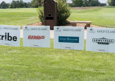 A row of sponsor signs is displayed on a grassy lawn at a golf event. The signs include logos and names such as "tribe," "EXODUS Moving & Storage," "Cline Williams Attorneys At Law," and "Armstrong hotel." Trees and a wooden box are visible in the background.