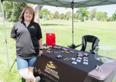 A woman stands smiling next to a table under a canopy at an outdoor event. The table displays various promotional items and is covered with a black tablecloth featuring the logo and name "Sprague Roofing & Exteriors." The background is a grassy area with trees.