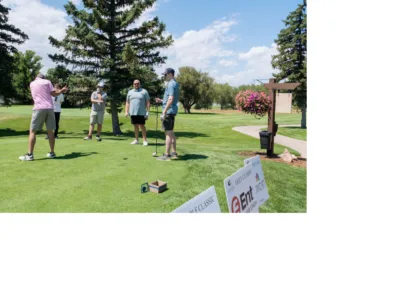 Four men stand on a golf course, engaging in conversation. One man is holding a golf club, preparing for a swing. A fifth person is seen in the background. The lush green course is complemented by trees and a flower basket. Signs for sponsors are displayed in the foreground.