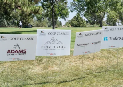 Four signs are on a grassy area near a tree, promoting various sponsors for a golf classic event. The sponsors listed are Adams Bank & Trust, Fire Tribe Video Content Creation, Coloradoan LocaliQ, and The Group.