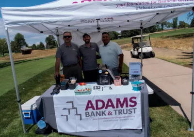 Three men stand behind a promotional booth for Adams Bank & Trust at an outdoor event. The booth features various branded items on the table, including drinks and snacks. A golf cart is visible in the background, and the scene appears to be set on a golf course.