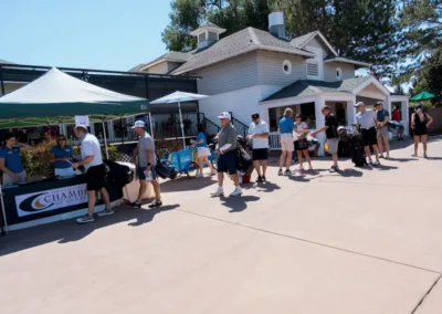 Participants stand in line outside a registration tent at a golf event. A banner on the table reads "Chamber of Commerce." People are wearing casual clothes and hats. The background shows a building and trees under a clear blue sky.