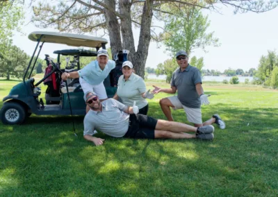 Four people pose cheerfully outdoors on a golf course near a golf cart. One person lies on the grass, while the other three stand next to or lean on the cart. Trees and a lake are visible in the background under a clear sky.