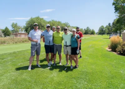 A group of six people standing on a golf course, dressed in casual golfing attire. They are smiling and posing with their golf clubs. The sky is clear and the grass is green, with trees in the background.