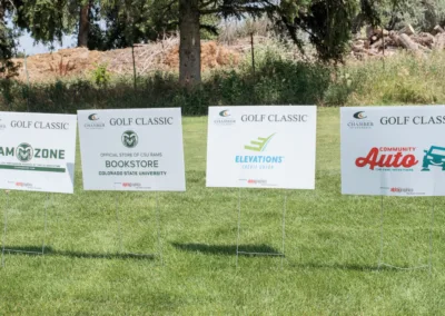 Four signs on a grassy area at a golf classic event display logos and names of sponsors: RAM ZONE, Colorado State University Bookstore, Elevations Credit Union, and Community Auto. Trees and greenery are visible in the background.