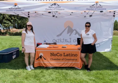 Two women stand in front of a canopy tent with "NoCo Latino Chamber of Commerce" signage. They are outdoors on a grassy area with trees in the background. A table covered with an orange tablecloth and promotional materials is centered under the tent.