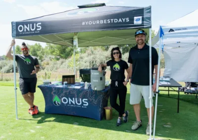 Three people stand under a tent with branding that reads "ONUS IV Hydration Bar" and "Your Best Days." There is a table with promotional materials and hydration products. The setting appears to be an outdoor event on a grassy area under a blue sky.