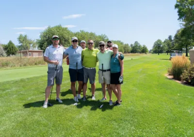 A group of six people stands on a golf course, smiling at the camera. They are wearing casual golf attire and hats. Some are holding golf clubs. The sunny day highlights green grass, trees, and a small building in the background.