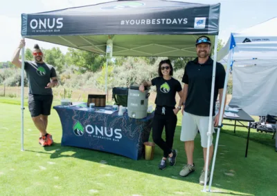 Three people standing under a tent on a grassy field. The tent has a banner that reads "ONUS IV Hydration Bar" and features a table with promotional items. One person is leaning on the table, another stands next to an IV drip, and the third person is holding a drink.