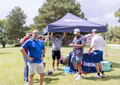 A group of people gather under and around a U.S. Bank tent set up on a grassy area with trees in the background. Some are wearing casual clothes, sunglasses, and caps. They appear to be engaged in conversation and looking at devices or papers.