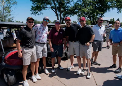 A group of seven men pose outside next to golf carts on a sunny day. They are dressed in various golfing attire, including polo shirts, shorts, and caps. Trees and a clear blue sky are visible in the background.