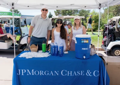 Three people stand behind a table with a blue cloth that reads "JP Morgan Chase & Co." The table holds water bottles, a jug, and several giveaway items. They are outdoors near golf carts, suggesting an event or tournament. Two of the people are wearing caps.