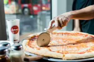 A person is cutting a large cheese pizza with a pizza cutter. A white cup with the logo "Anthony's" is on the table next to shakers of Parmesan cheese and red pepper flakes. The background shows a blurred view of a window and a red vehicle outside.