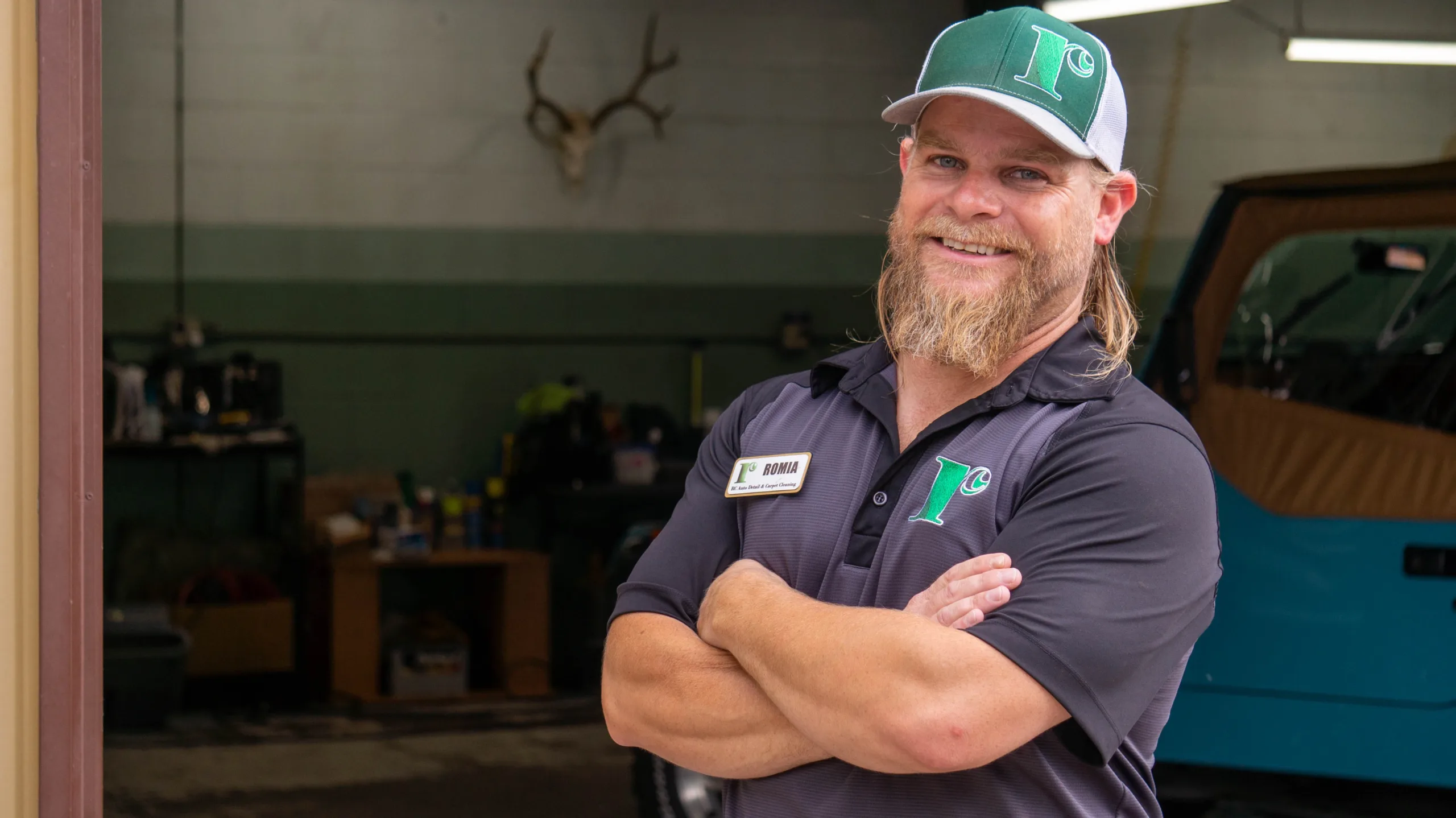 A man with a beard and a white cap stands confidently with arms crossed in front of a service menu for an auto detailing and carpet cleaning business. The menu lists various detailing services and add-on items with prices. Promotional posters are visible on the wall.