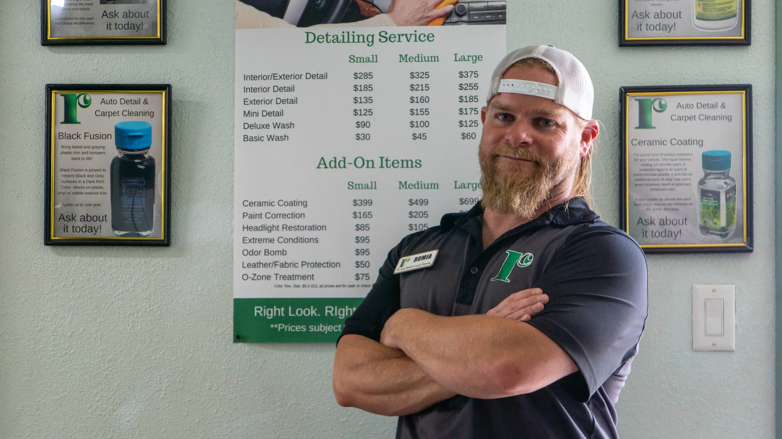 A man with a beard and a white cap stands confidently with arms crossed in front of a service menu for an auto detailing and carpet cleaning business. The menu lists various detailing services and add-on items with prices. Promotional posters are visible on the wall.