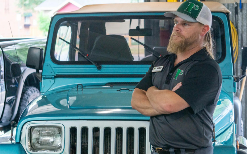 A bearded man wearing a green and white cap, a black shirt, and a name tag stands with his arms crossed in front of a blue Jeep inside a garage.