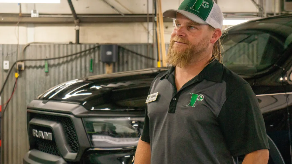 A person with a blonde beard and long hair, wearing a black polo shirt with a green logo and nametag, stands with folded arms in front of a truck inside a garage.