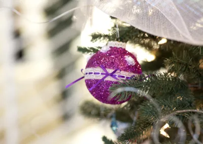 A sparkly purple Christmas ornament with white lace and ribbon details hangs on a green Christmas tree branch. The background is softly blurred, highlighting the festive decoration.