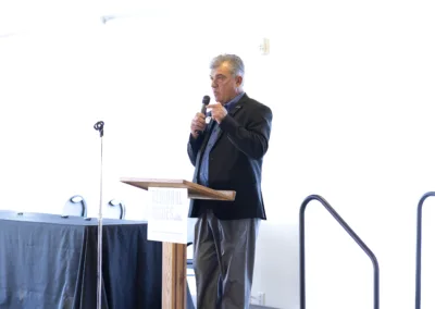 A man in a suit speaks into a microphone while standing at a wooden podium with a sign that reads "Regional Issues." The room is bright and minimally decorated, with a table and chairs visible in the background.