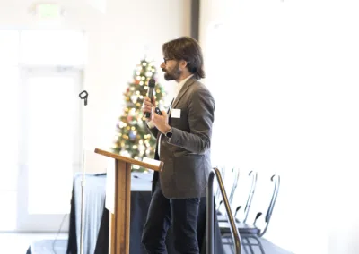 A man with a beard and glasses is giving a presentation at a podium in a bright room. He holds a microphone and gestures with his hand. A Christmas tree with lights is seen in the background. Empty chairs are lined up against the wall.