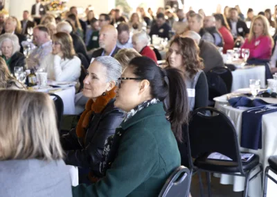 A diverse group of people attentively seated at round tables during a formal event, with some taking notes. Tables are set with glassware and documents, and attendees are focused on a speaker not visible in the image.