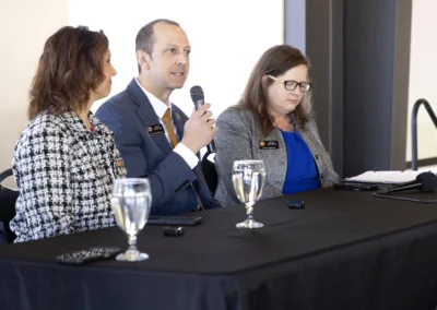 Three people are seated at a table with microphones and water glasses. The person in the center is speaking into a microphone. They appear to be at a panel discussion or conference in a well-lit room.