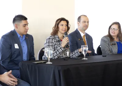 A group of four individuals is seated at a table with a black tablecloth. One woman is speaking into a microphone. They each have glasses of water in front of them. All are dressed in business attire, engaged in a discussion.