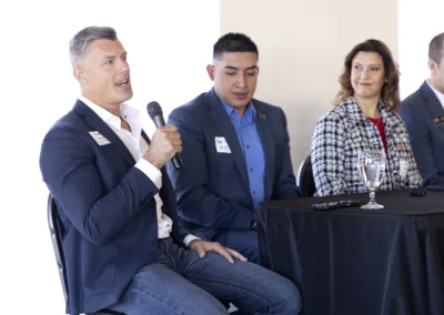 A man in a blue suit holds a microphone while speaking at a panel discussion. Three other panelists, two men and a woman, sit beside him at a table covered with a black cloth. A name tag is visible on each participant's jacket.