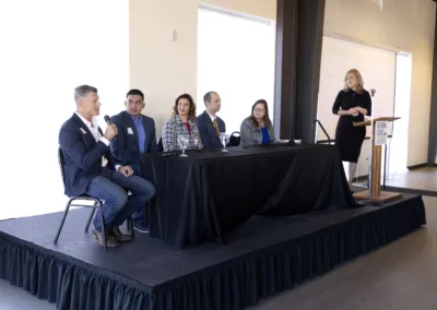 A panel of five people sit at a long table on a stage, engaged in discussion. One man holds a microphone. A woman stands at a podium to the right. They are in a well-lit room with large windows.