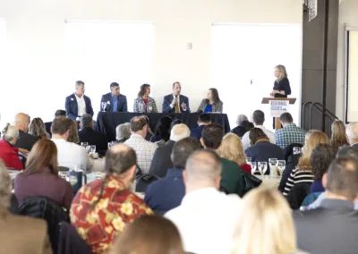 A panel of five people sits at a long table on a stage, speaking at a conference. The audience is seated in rows facing the panel. A woman stands at a podium labeled "Regional Issues.