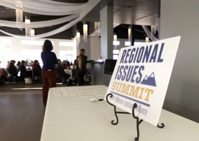 A conference room with people gathered at tables. In the foreground, a sign reads "Regional Issues Summit" on a stand, placed on a table with name tags. Draped fabric and pendant lights hang from the ceiling.