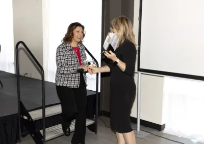 Two women stand near a podium. One, in a black dress, presents an award to the other, wearing a black and white patterned jacket and red top. They smile and shake hands, with a stage and screen in the background.