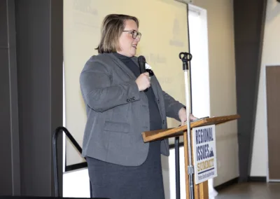 A person wearing glasses and a gray suit stands at a podium with a microphone, speaking at the Regional Issues Summit. A projection screen is visible in the background.