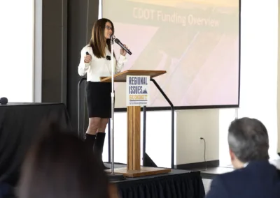 A woman stands at a podium with a microphone, presenting slides on "CDOT Funding Overview" at a "Regional Issues Summit." She wears a white blouse and black skirt, with a screen displaying a presentation in the background. Audience members listen attentively.