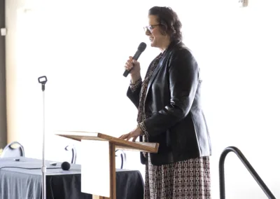 A person in a patterned dress and leather jacket speaks into a microphone while standing at a wooden podium. Several chairs and a microphone stand are visible in the background.