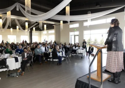 A woman speaks at a podium to a seated audience in a large event hall. The room features draped fabric from the ceiling and large windows. Attendees sit at round tables, engaging with the presentation.