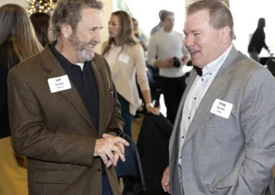 Two men in conversation at an indoor event. One wears a brown jacket and holds glasses, while the other wears a gray suit. Both have name tags. A small crowd is in the background, and a decorated tree is partially visible.