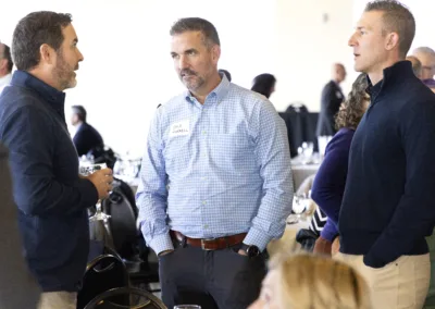 Three men are engaged in conversation at an indoor event. They are wearing casual business attire, and one has a name tag. In the background, people are seated at tables with white tablecloths.