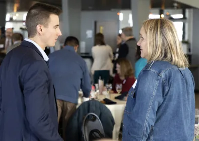 Two people, a man in a suit and a woman in a denim jacket, engage in conversation at a social event. They stand near tables where other attendees are seated and talking, creating a lively atmosphere.