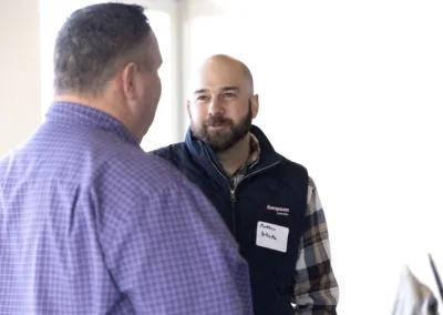 Two men are having a conversation indoors. The man facing the camera is wearing a dark vest over a plaid shirt and a name tag. The other man has short hair and is wearing a purple checkered shirt. The background is bright and slightly blurred.