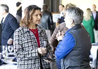 Two women are engaged in conversation at an indoor event. One wears a black and white jacket, and the other a blue shirt and gray vest. People are mingling in the background with tables and chairs around them.