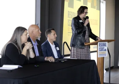 Four individuals are participating in a panel discussion at a "Regional Issues Summit." One person is speaking at a podium, while the others sit at a table listening. A screen in the background displays the event name.