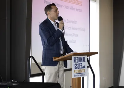 A man in a checkered blazer speaks into a microphone at a podium during a conference. The podium has a sign reading "Regional Issues Summit." A screen behind him displays text.